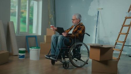Photo for An elderly disabled man moves in a wheelchair plans renovation using a digital tablet. A pensioner checks building materials. Room with window, ladder, cardboard boxes, wallpaper rolls, paint bucket - Royalty Free Image