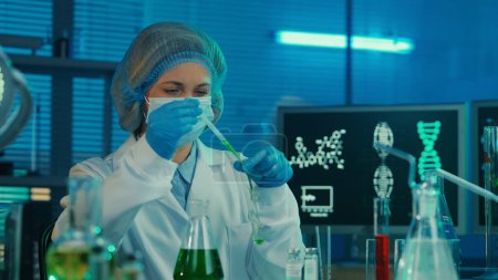 Téléchargez les photos : Une scientifique pipette un liquide vert dans un tube à essai en verre. Une femme vêtue d'une blouse médicale blanche, d'un bonnet, de gants bleus et d'un masque dans un laboratoire avec éprouvettes, flacons et écrans d'ordinateur en bleu - en image libre de droit