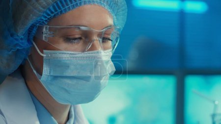 Téléchargez les photos : Portrait d'une femme médecin dans un masque médical, lunettes, robe blanche et bonnet bleu regarde vers le bas. Scientifique ou chercheuse posant sur fond d'un laboratoire ou d'un hôpital moderne au bleu - en image libre de droit