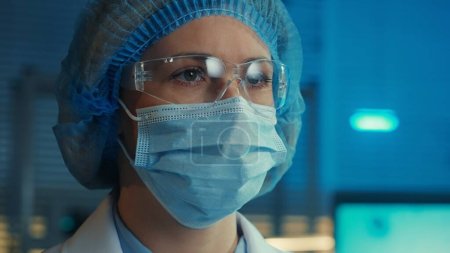 Photo for Portrait of a woman doctor in a medical mask, goggles, white gown and blue bonnet looks away. Female scientist or researcher posing against the backdrop of a modern laboratory or hospital with blue - Royalty Free Image