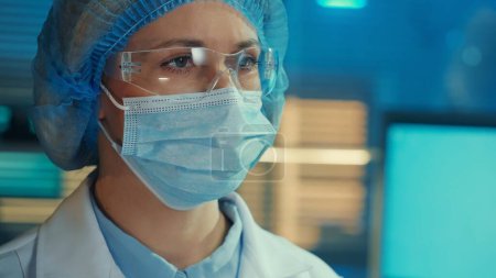 Téléchargez les photos : Portrait d'une femme médecin dans un masque médical, lunettes, robe blanche et bonnet bleu regarde loin. Scientifique ou chercheuse posant sur fond d'un laboratoire ou d'un hôpital moderne au bleu - en image libre de droit