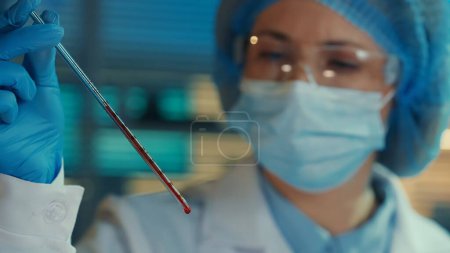 Photo for A woman carefully examines a glass pipette filled with red liquid. Female doctor or researcher in goggles, bonnet, mask, white gown and blue gloves in biochemical laboratory or hospital. Blue light - Royalty Free Image