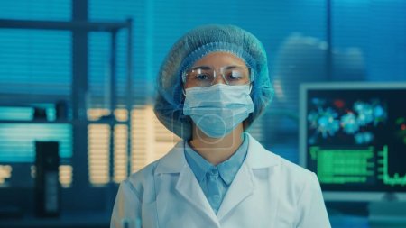 Photo for Portrait of a woman doctor in a medical mask, goggles, white gown and blue bonnet looks directly into the camera. Female scientist or researcher posing against the backdrop of a modern laboratory or - Royalty Free Image