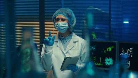 Téléchargez les photos : Femme médecin examine les flacons en verre, les flacons, les éprouvettes et entre les données dans la tablette numérique. Scientifique en blouse blanche, bonnet, lunettes, masque et gants dans un laboratoire ou un hôpital avec lumière bleue - en image libre de droit