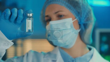 Photo for Portrait of a woman in a white gown, medical mask, bonnet and blue gloves holds a glass vial with a vaccine in her hand. A female doctor examines the vial on the background of a laboratory or hospital - Royalty Free Image