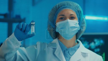 Photo for Portrait of a woman in a white gown, medical mask, bonnet and blue gloves holds a glass vial with a vaccine in her hand. A female doctor looks into the camera on the background of a laboratory or - Royalty Free Image