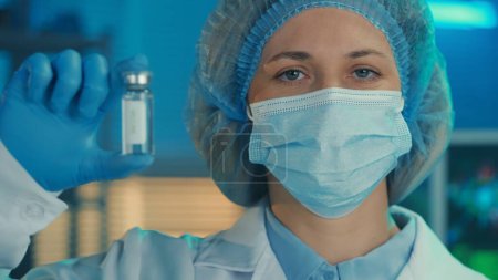 Foto de Retrato de una mujer vestida con una bata blanca, máscara médica, gorro y guantes azules sostiene un frasco de vidrio con una vacuna o un medicamento en la mano. Una doctora mira a la cámara en el fondo de un - Imagen libre de derechos