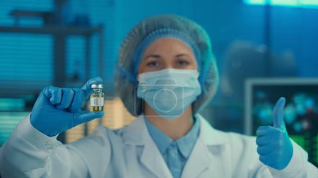 Téléchargez les photos : Une femme tient dans sa main un flacon en verre du vaccin contre le virus CoV-2 du SRAS et montre un pouce en l'air. Portrait d'une femme médecin en robe blanche, masque médical, bonnet et gants bleus sur le fond d'une - en image libre de droit