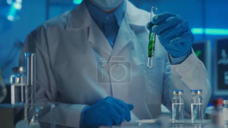 Téléchargez les photos : Un médecin masculin vêtu d'un manteau blanc et de gants bleus tient dans sa main une éprouvette en verre avec une substance verte se dissolvant dans un liquide transparent. Etude des échantillons, réaction chimique. Biochimique moderne - en image libre de droit
