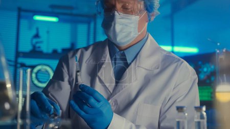 Foto de Un hombre con una bata blanca, máscara médica, gafas y guantes azules sostiene una jeringa con una vacuna o un medicamento en las manos. Laboratorio de investigación bioquímica moderna, de cerca. Desarrollo de una innovación - Imagen libre de derechos