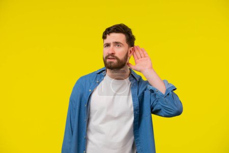 Photo for Young, serious, blond man in a blue shirt on a yellow background, holding an ear to listen better, what youre saying. Photo Shoot. - Royalty Free Image