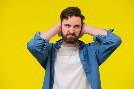 Photo for Shocked upset middle aged caucasian guy with beard covers his ears and does not want to hear, isolated on yellow background, panorama. Man feels depressed, suffers from noise and stress, copy space - Royalty Free Image