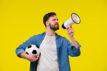 Photo for Very happy cheerful man with beard in striped t-shirt blowing in big bullhorn holding football ball, celebrating championship beginning. Indoor studio shot isolated on yellow background - Royalty Free Image