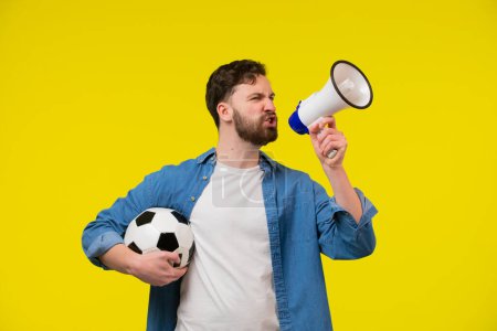 Photo for Very happy cheerful man with beard in striped t-shirt blowing in big bullhorn holding football ball, celebrating championship beginning. Indoor studio shot isolated on yellow background - Royalty Free Image