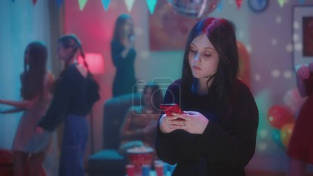 Photo for Goth girl watching video on phone, typing a message, checking social media while her friends are having fun at the party dancing. Single girl. The room is smartly decorated for a house party with a - Royalty Free Image