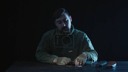Photo for A soldier with a Ukrainian flag on his chest unloads a Kalashnikov assault rifle magazine. A man sits at a wooden table on a black background. Close up. Army training - Royalty Free Image