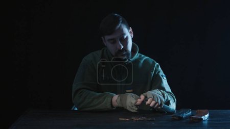Foto de Un soldado carga un cargador de armas de fuego con cartuchos tendidos frente a él en una mesa de madera. Primer plano de un soldado ucraniano sentado en una mesa sobre un fondo negro. Entrenamiento militar - Imagen libre de derechos