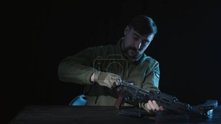 Photo for A Ukrainian soldier assembles a Kalashnikov assault rifle. Close-up of a Ukrainian soldier sitting at a wooden table with a rifle in his hands on a black background. Preparation for the army - Royalty Free Image