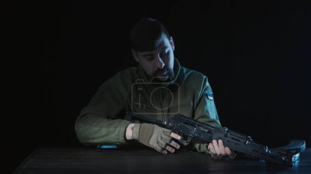Photo for A soldier of the armed forces of Ukraine sits at a table with a Kalashnikov assault rifle in his hands. Close up of a soldier on a black background. Preparation for the army, development of skills - Royalty Free Image