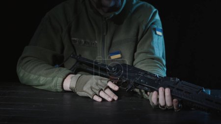 Photo for A soldier of the armed forces of Ukraine sits at a table with a Kalashnikov assault rifle in his hands. Rifle in the hands of a soldier on a wooden table close up - Royalty Free Image