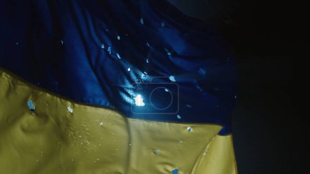 Foto de El desarrollo de la bandera azul amarilla de Ucrania sobre un fondo negro iluminado por un foco. Bandera de Ucrania cubierta con agujeros de bala de cerca. Apoyar a Ucrania - Imagen libre de derechos