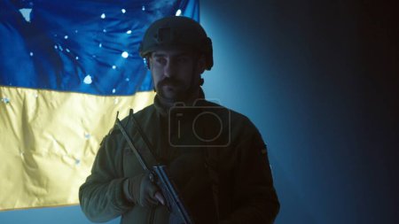 Foto de Un primer plano de un soldado ucraniano usando un casco y sosteniendo un rifle de asalto Kalashnikov. Un soldado se levanta en el fondo de la bandera de Ucrania plagada de agujeros de bala en la oscuridad, en la - Imagen libre de derechos