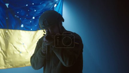 Foto de Un soldado de las fuerzas armadas de Ucrania apunta a un rifle de asalto Kalashnikov. Un soldado en un casco se levanta contra el fondo de la bandera de Ucrania en los rayos de un foco. De cerca. El - Imagen libre de derechos