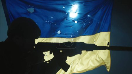 Photo for Profile portrait of a silhouette of a Ukrainian soldier aiming a weapon in the dark. Close up of the silhouette against the background of the Ukrainian flag, covered with bullet holes, lit by a - Royalty Free Image