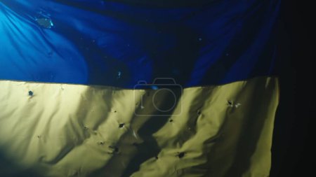 Téléchargez les photos : Le drapeau bleu jaune en développement de l'Ukraine sur un fond noir illuminé par un projecteur. Drapeau de l'Ukraine recouvert de trous de balle fermer. Stand avec l'Ukraine - en image libre de droit