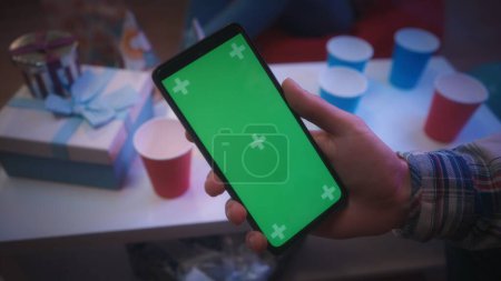 Close up of a mans hand holding a smartphone with a green screen on the background of a table with paper red, blue cups and gifts. Uses the phone to browse the Internet, photo, video. The concept of