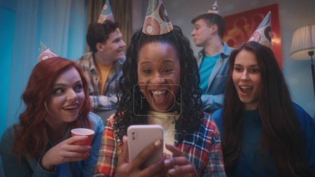 Téléchargez les photos : Un groupe de jeunes en chapeaux de fête lors d'une fête d'anniversaire à la maison. Les copines, assises sur le canapé, sont surprises, choquées et regardent joyeusement le téléphone, tenu par une Afro-Américaine - en image libre de droit