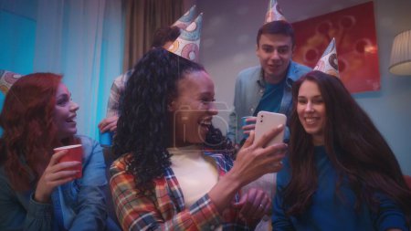 Téléchargez les photos : Une femme afro-américaine montre le téléphone à ses amis, ils regardent émotionnellement des photos, des vidéos. Un groupe d'amis dans des chapeaux de fête dans une pièce décorée pour une fête, avec des réflexions de balle disco sur le - en image libre de droit