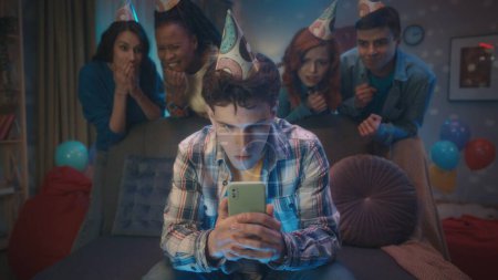 Téléchargez les photos : Un gars joue à un jeu en ligne sur son téléphone, ses amis le regardent se tenir intensément derrière lui. Jeux, divertissement lors de fêtes à domicile - en image libre de droit