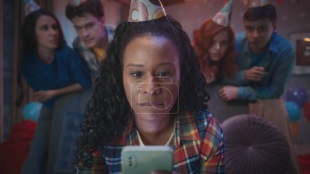 Téléchargez les photos : Une Afro-Américaine joue à un jeu en ligne sur son téléphone, se mordant les lèvres à cause de la tension. Les amies se tiennent derrière elle et s'inquiètent pour elle. Un groupe de jeunes en chapeaux de fête à un - en image libre de droit