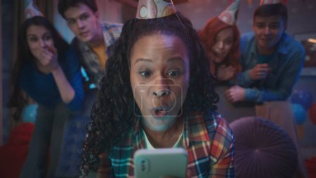 Foto de Una chica afroamericana con un sombrero festivo se ve sorprendida, conmocionada por el teléfono. Las amigas están detrás de ella. Retrato de cerca de la cara sorprendida de una chica sentada en un sofá en un - Imagen libre de derechos