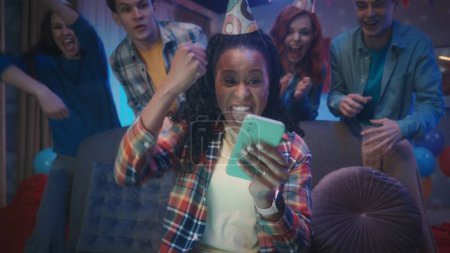 Téléchargez les photos : Une fille afro-américaine joue à un jeu en ligne sur son téléphone pendant que ses amis la regardent de derrière elle. La jeune fille gagne, agitant la main dans un geste de victoire. Soirée maison. Jeux - en image libre de droit