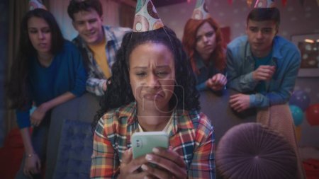 Téléchargez les photos : Un groupe de jeunes gens en chapeaux de fête regardent leur ami afro-américain jouer à un jeu en ligne sur son téléphone. La fille perd et se fâche, ses amis lui remontent le moral. Divers jeux et - en image libre de droit