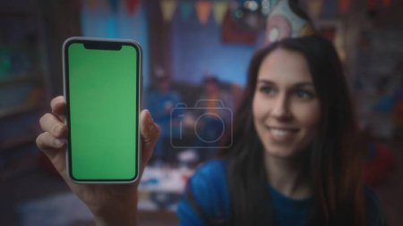 Téléchargez les photos : Une fille dans un chapeau de fête montre un smartphone avec un écran vert, sourit. Gros plan du smartphone écran vert sur un fond flou d'une fête, dans une pièce décorée d'une boule disco, drapeaux. Technologie - en image libre de droit