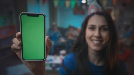 Foto de Una chica con un sombrero festivo muestra un teléfono inteligente con una pantalla verde, sonrisas. Primer plano del smartphone de pantalla verde sobre un fondo borroso de una fiesta, en una habitación decorada con una bola de discoteca, banderas. Tecnología - Imagen libre de derechos