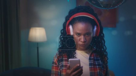 Foto de Chica afroamericana en auriculares inalámbricos rojos pasa a través de una lista de reproducción en el teléfono. Retrato de cerca de una chica afroamericana en una habitación, cuyo interior está decorado con un espejo - Imagen libre de derechos