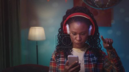 Foto de Una chica afroamericana con auriculares inalámbricos rojos hojea una lista de reproducción en su teléfono con su dedo índice. Retrato de cerca de una chica afroamericana en una habitación, cuyo interior es - Imagen libre de derechos