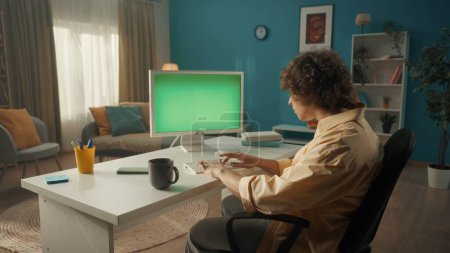 Foto de Un joven rizado está trabajando en una computadora con una pantalla verde. Un hombre se sienta en una mesa con una computadora, y hay una taza de café a su lado. Vista trasera. El concepto de tecnología, comunicación - Imagen libre de derechos