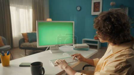 Foto de Un joven rizado trabaja en una computadora con una pantalla verde de cerca. Un hombre se sienta en una mesa con una computadora, y hay una taza de café a su lado. Vista trasera. El concepto de tecnología, comunicación - Imagen libre de derechos