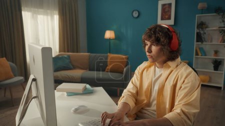 Téléchargez les photos : Un jeune homme sérieux et concentré dans un casque sans fil rouge est assis sur un ordinateur. Un jeune homme joue à un jeu en ligne, travaille, regarde des vidéos, écoute de la musique. Gros plan - en image libre de droit