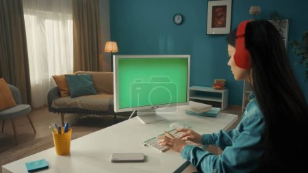 Téléchargez les photos : Une jeune femme asiatique travaille sur un ordinateur avec un écran vert de près. Une brune aux cheveux longs s'assoit à une table en casque sans fil rouge. Vue latérale. Le concept de technologie, de communication - en image libre de droit