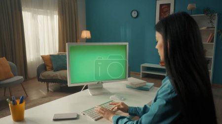 Foto de Una joven asiática trabaja en una computadora con una pantalla verde de cerca. Una morena de pelo largo se sienta en una mesa de estar. Vista lateral. El concepto de tecnología, comunicación, conexión - Imagen libre de derechos