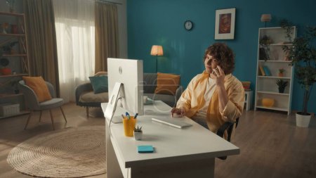 Téléchargez les photos : Un jeune bouclé est assis à un ordinateur avec un smartphone à l'oreille. Un homme parle au téléphone pendant qu'il travaille sur l'ordinateur. Le concept de technologie, de communication, de connexion - en image libre de droit