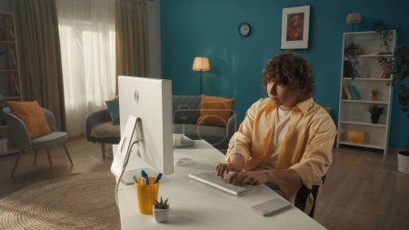 Foto de Un joven está sentado frente a una computadora en una acogedora sala de estar. Un hombre está escribiendo en el teclado, escribiendo correos electrónicos, mensajes, chats, navegar por Internet, freelancer. El concepto de tecnología - Imagen libre de derechos