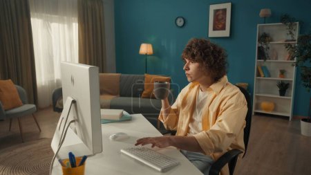Foto de Un joven rizado está sentado en una computadora. En una mano, un joven sostiene una taza de café, la otra está escribiendo en el teclado. Un hombre escribe correo electrónico, mensajes, chats, trabaja en Internet, freelancer - Imagen libre de derechos