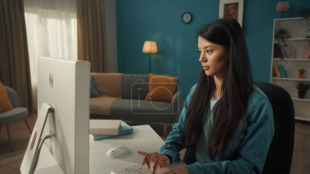 Foto de Una mujer asiática se sienta en una computadora en una acogedora sala de estar. Mujer joven trabajando, escribiendo en el teclado, escribiendo correos electrónicos, mensajes, chats, navegando por Internet, freelancer - Imagen libre de derechos